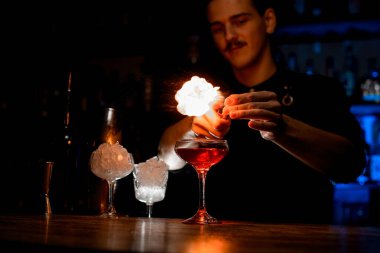 Barmen uzun saplı bir kokteyl bardağının üstünde ateş yakıyor. Yanında da buzlu iki bardak var.