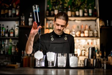 Becerikli barmen, barın tezgahında duran bardaklara şişeden mavi bir içki doldurur. Her birinin yanında ezilmiş buzlu bir bardak vardır.