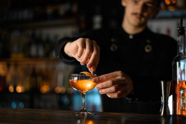 Barmen, bulanık bir arka planda kahverengi bir kokteyl ile dolu bir bardağa küçük bir mandalla bir parça tat ekliyor.