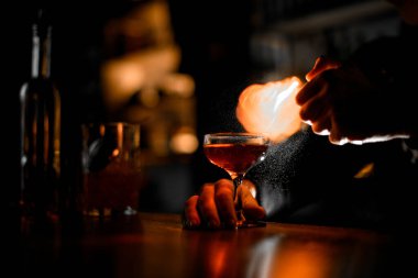 Barmenlerin elleri bardağı bir kokteyl ile tutar ve koyu bulanık arka planda alev spreyi sıkarlar.