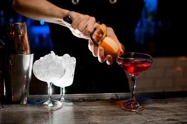 Glass with a cocktail stands on the bar counter, glasses with crushed ice are nearby, and the bartenders hands cut off a piece of zest next to the cocktail