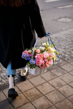 Siyah beyaz ayakkabılı ve açık mavi pantolonlu bir kız elinde çiçeklerle elinde bir sepetle yürüyor.