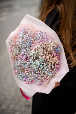 Ellerinde mavi-pembe-beyaz-bej renkte çiçekler var. Gün boyunca siyah ceketli bir kızın elinde.