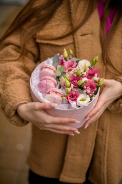 Açık kahverengi ceketli, karton kutulu bir kız. Şefkat çiçekleri ve makaronlar şehrin caddesinde duruyor.