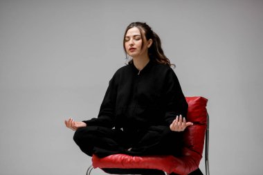 Siyah takım elbiseli genç bir kadın meditasyon yapıyor. Kırmızı deri koltukta oturuyor. Nilüfer pozisyonunda metal bacakları var.