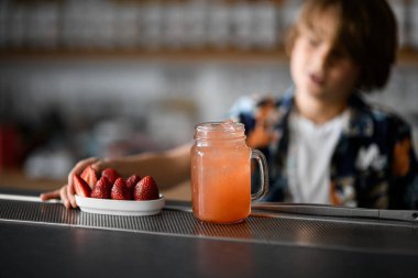 Focus on a glass jar with a handle filled with a pink fruit cocktail next to a saucer with strawberries, a boy as a bartender standing on a blurred background clipart