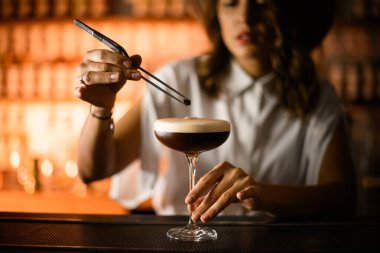 Barmenin cımbızla süslediği kahverengi köpüklü kahve kokteyli olan uzun saplı bardağın ön planına odaklan.