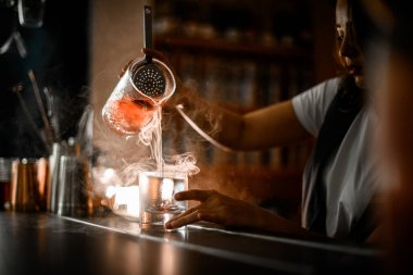 Female bartender pours a brown cocktail with ice from one glass to another, straining the drink through a sieve, smoke spreads near the glasses clipart