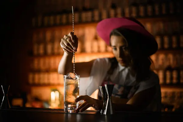 Stock image Female bartender stirs a long bar spoon with a clear cocktail in a tall glass with three ice balls on a blurred background