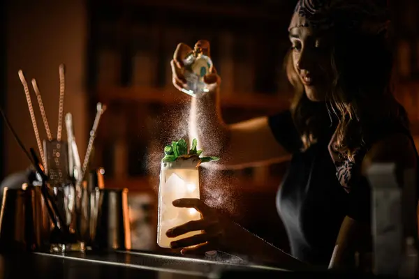 Stock image Happy pretty woman bartender spraying a glass with a cocktail covered with condensation, splashes of spray flying, illuminated by a blurred background