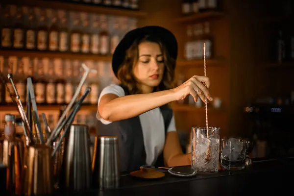 Stock image Female bartender stirs ice in a glass with a long bar spoon to prepare a cocktail against the background of an illuminated bar showcase