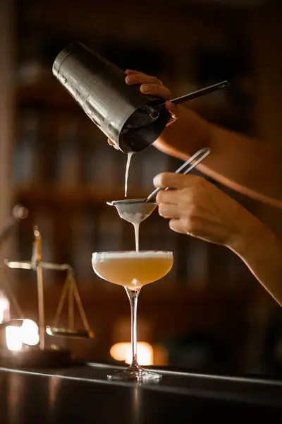 stock image Female bartender finishes straining through two sieves a frothy champagne-colored cocktail into a stemmed glass standing on a bar counter on a blurred background