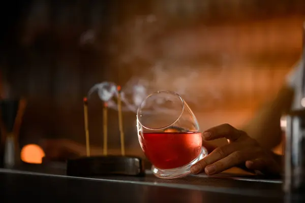 stock image Female bartender rubs an unusually shaped tumbler with a convex bottom, filled with a pink cocktail with ice, incense sticks are smoking nearby