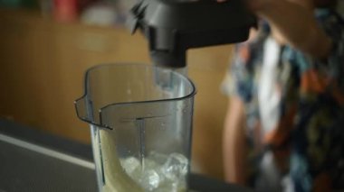 Temiz bir blender buz ve muz parçasıyla dolu bir tezgahın üzerinde duruyor. Blender odak noktasındayken parlak renkli gömlekli bir kişi arka planda odak dışı kalıyor..