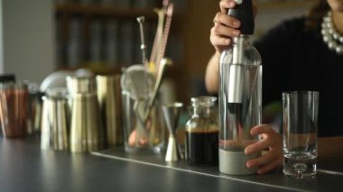Bir barmen, karbonatlı suyla birlikte cam bir şişe kullanarak içki hazırlar. Barmen suyu dikkatlice uzun bir bardağa doldurur. Tezgahın arkasında çeşitli kokteyl aletleri ve şişeler var.