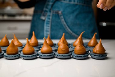 Beyaz mermer bir masada kahverengi kremalı mavi gri badem kurabiyesi ve mutfakta mavi kot pantolon giyen pasta şefi.