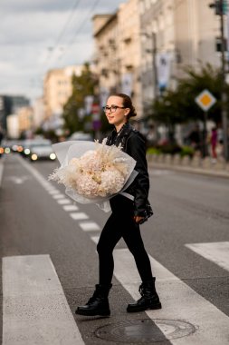 Gözlüklü, gülümseyen güzel bir kız, elinde kocaman, yumuşacık bir buket çiçekle sokağa çıkıyor.