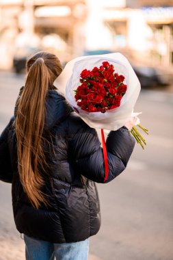 Koyu renk saçlı, siyah elbiseli ve gözlüklü genç bir kadın elinde caddede bir buket kırmızı gül tutuyor.