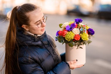 Uzun siyah saçlı ve gözlüklü tatlı kız bulanık bir arka planda duruyor çok renkli çiçeklerle