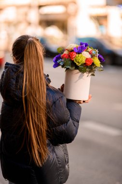 Genç kız elinde, şehir sokaklarının bulanık arka planında, karton bir kutuda çok renkli güzel bir çiçek tutuyor.