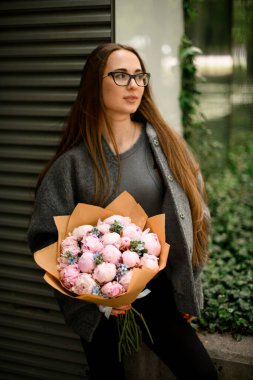 Gözlüklü, uzun kahverengi saçlı, gri, kalın elbiseli, şefkat dolu pembe çiçekli genç bir kız.