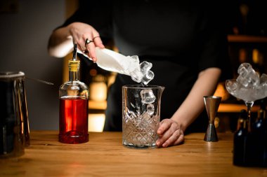 Female bartender hands scooping ice cubes into a glass for mixing cocktails, a geyser bottle filled with brown drink stands next to it clipart