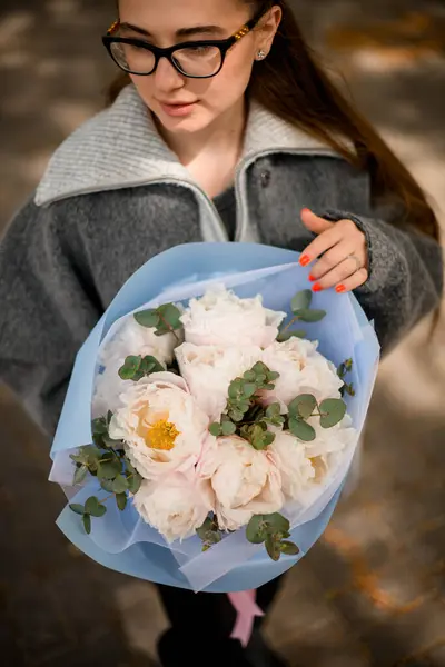 stock image Innocent girl in a grey cloth with a big bouquet of chic flowers in blue wrapper stands on a city street