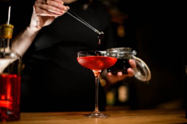 Üzerinde kahverengi kokteyl dolu uzun saplı şarap bardağına odaklan. Hangi barmen kadın elinde cımbızla kokteyl damlayan böğürtlen tutar?