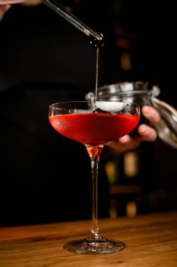 Kahverengi bir kokteyl ile doldurulmuş saplı bir şarap kadehinin yakın çekim odağı. Tam üzerinde bir bayan barmen elinde cımbızla bir dut tutuyor.