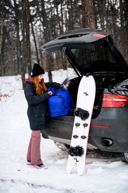 Young girl takes out backpack and snowboard from car during stop in winter forest. Winter trip by car. Winter snowboarding season clipart