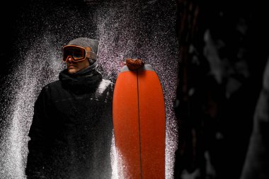 Elinde snowboard tutan koyu renk spor giysili genç adam karanlıkta karın altında duruyor. Yan görüş. Metin için boşluk