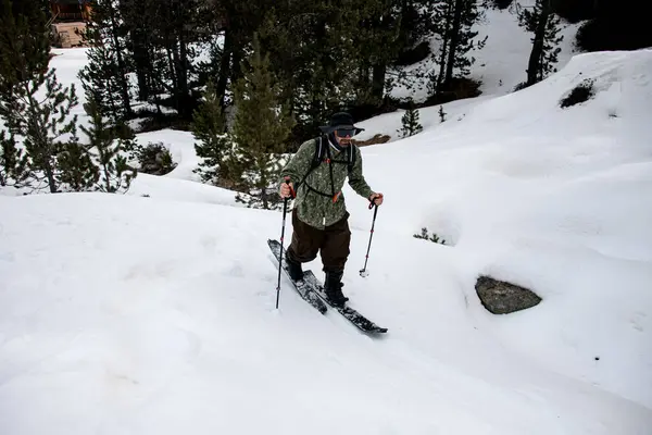 stock image Skier in black glasses and a wide-brimmed hat skis up a snowy slope against the background of tall conifers