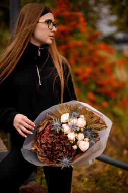 Çikolata ortancalarından, beyaz güllerden ve kraft kağıdında çan çiçeklerinden oluşan kocaman bir sonbahar buketi tutan güzel bir kadın.