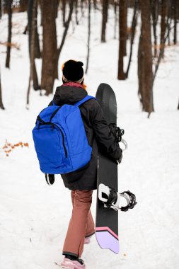 Karlı bir ormanın arka planına karşı omuzlarında büyük bir sırt çantası taşıyan, snowboard 'la yürüyen bir kadının fotoğrafı.