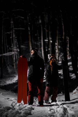 Erkek snowboardcu yanında bir bölme tahtası tutuyor ve bir kadın elinde bir snowboard tutuyor ve farklı yönlere bakıyor.