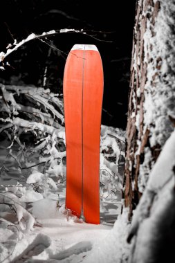 Red splitboard stands slightly submerged in snow next to tree covered with snow in the dark with low lighting clipart