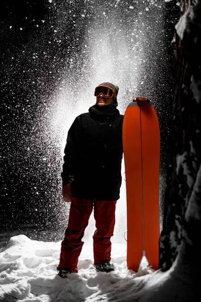 Kar gözlüklü genç erkek snowboardcu, yandan ona bakıyor, arkasında toz kar arkaplanı duruyor.