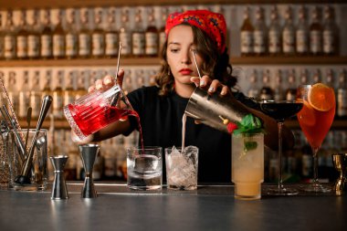 Kadın barmen, bulanık bir arkaplanda buzlu iki kadehten iki bardağa nasıl farklı kokteyller koyduğuna konsantre oluyor.