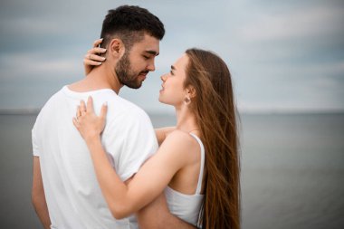 Genç, yakışıklı bir adam ve bir kadın birbirlerine sarılıyorlar, gülümsüyorlar ve birbirlerinin gözlerine bakıyorlar. Deniz kıyısında güzel bir tatil.