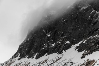 Kalın bir sis tabakasıyla kaplanmış dik bir kayanın yan görüntüsü. Bir kısmı karla kaplı.