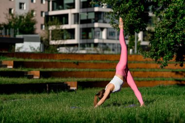 Genç, sıska bir kadın köpek yogasına bakıyor. Bir bacağı yukarı doğru uzanmış, şehrin ortasında yeşil bir çimenlikte.