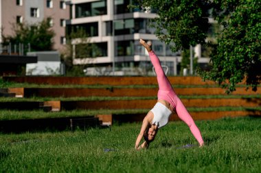 Genç, narin bir kadın, bir ayağı çimlerin üzerinde dikilip ellerini bulanık bir arka planda dinlendirirken havada ikiye ayrılıyor.