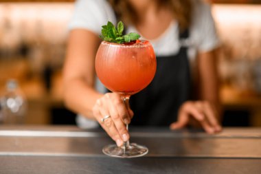 Soğuk turuncu bir kokteyl ve arka planda bulanık bir nane yaprağı olan uzun bir bardağın yakın çekimi beyaz tişörtlü bir bayan barmenin siluetini gösteriyor.