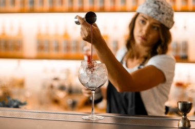 Ön plana odaklan, uzun bir bardak bir bar tezgahında buzla doludur, dişi bir barmen bir zencefilden koyu kahverengi bir içecek doldurur, bardağın üzerinde, bulanık bir şekilde,