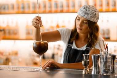 Kadın barmen, barın üstünde duran yüksek saplı bir bardakta buzla kahverengi bir kokteyl karıştırıyor. Kokteylli cama odaklan. Bulanık arkaplan