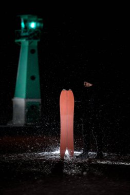 Young man looks to the side, holding a red splitboard standing on the sand on the background of a glowing green pendulum, small snowflakes are visible on the background of a lantern clipart