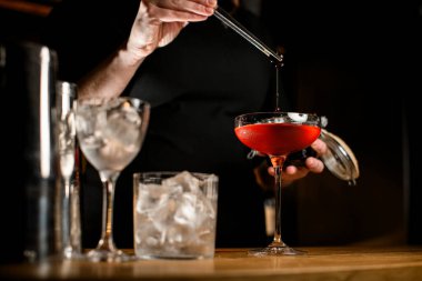 Üzerinde kahverengi bir kokteyl bulunan uzun saplı şarap bardağına odaklan. Hangi barmen kadın elinde cımbızla meyve suyu tutarak meyve suyunu doğrudan kokteylin içine sıkıyor?