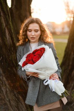 Pretty young woman holding a bouquet of red roses in white wrapping paper, leaning against a tree trunk against a sunset background clipart