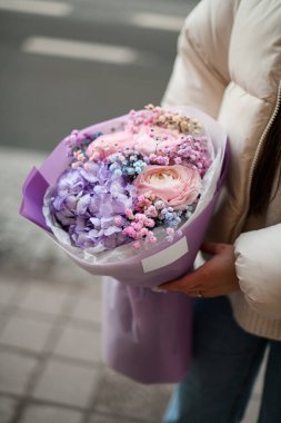 Blue, pink, white and beige flowers in the hands of a girl who stands on a city street in a a light puffy jacket during the day clipart