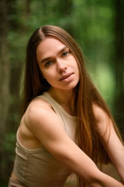 Uzun, dalgalı saçlı güzel bir kadının portresi sakince yeşilliğin bulanık arka planında kameraya bakıyor.